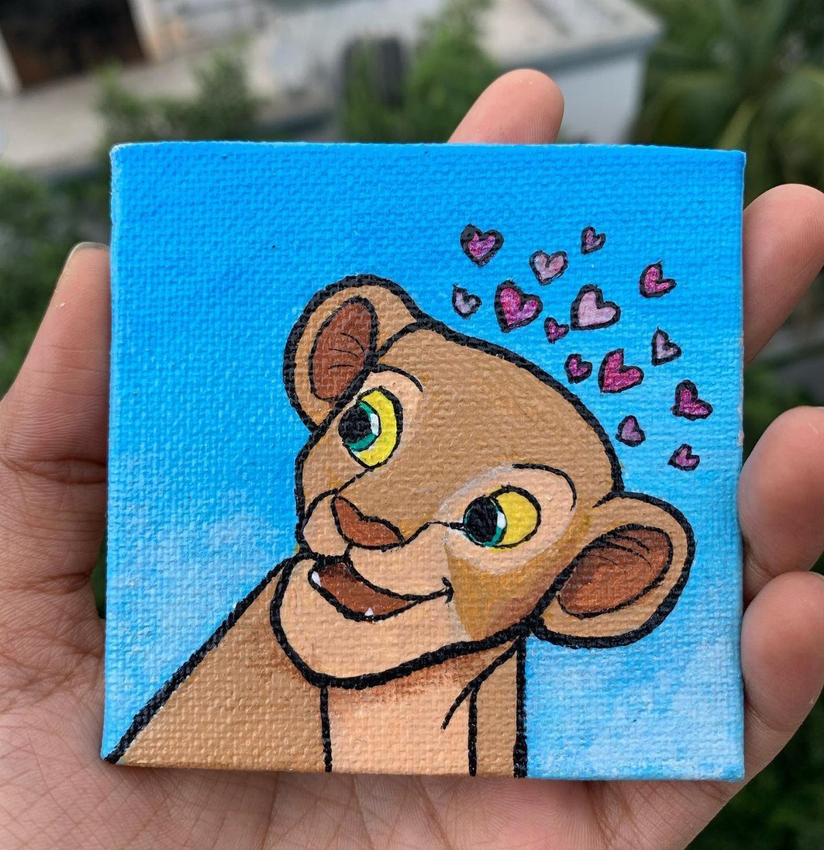 A hand holding a small square canvas painted with a cartoon lion cub and pink hearts.