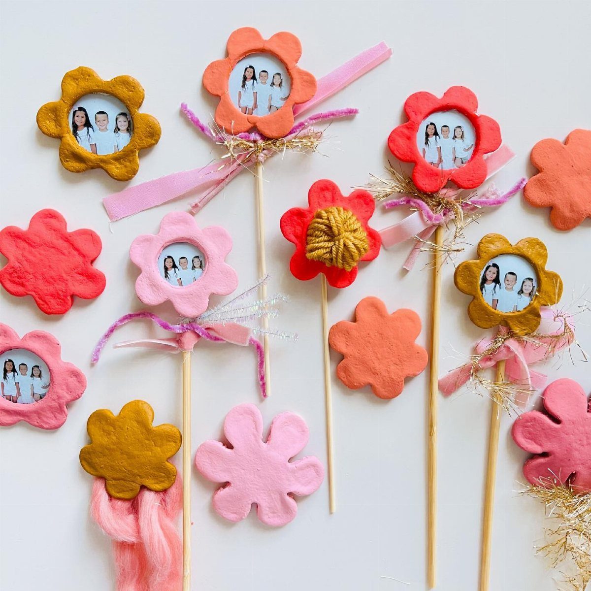 Colorful flower-shaped clay magnets with small photos and decorative ribbons on wooden sticks.