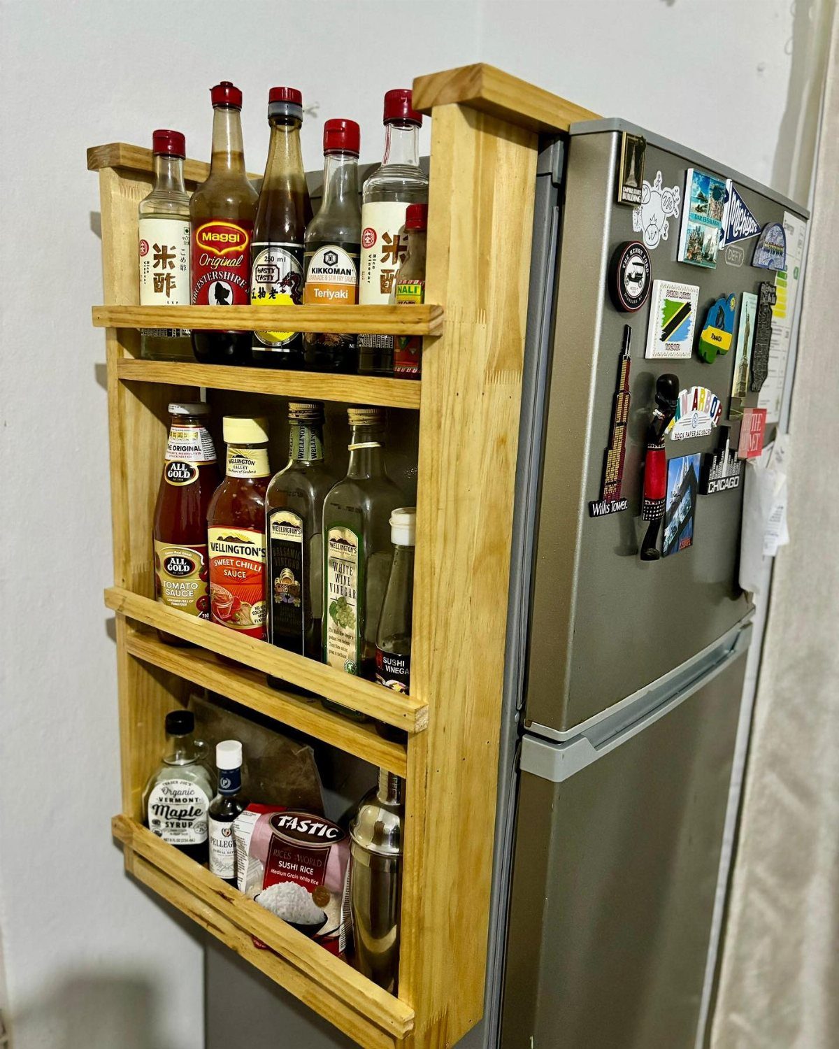 Wooden shallow spice shelves mounted to the side of a stainless steel refrigerator filled with bottles.