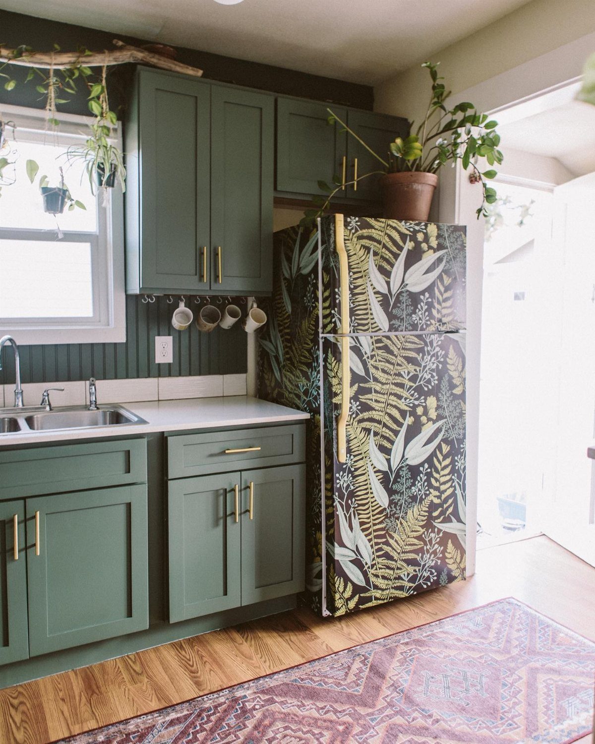 A refrigerator decorated with dark botanical leaf pattern wallpaper in a green kitchen.