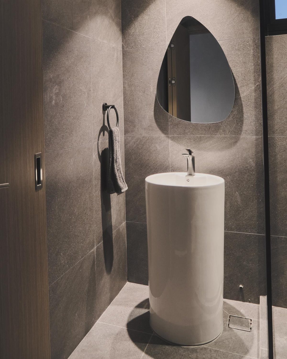 A white cylindrical pedestal sink positioned against grey tiled walls with an organic shaped mirror.