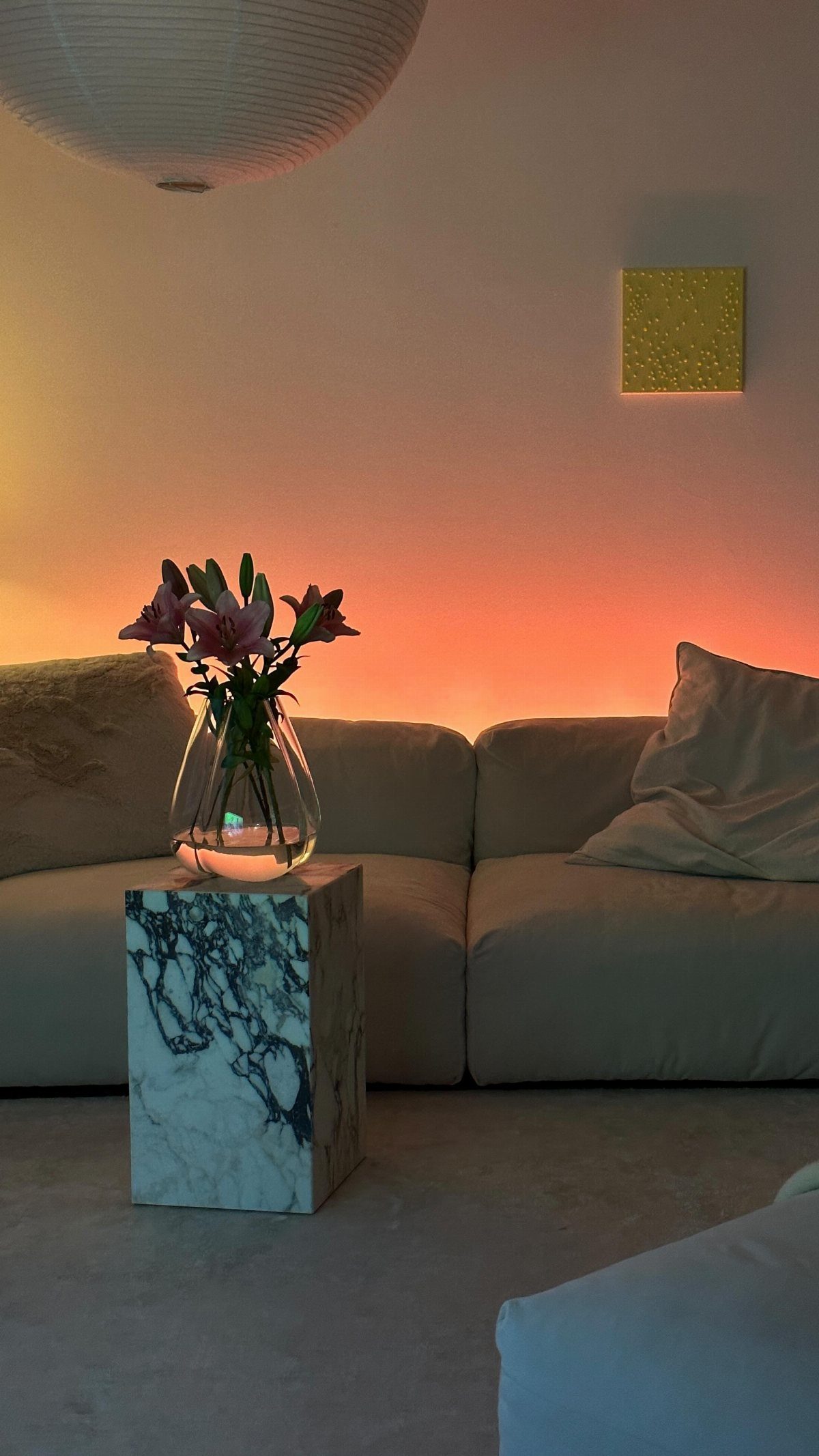 Pink lilies in a glass vase on a marble pedestal with warm orange ambient lighting.
