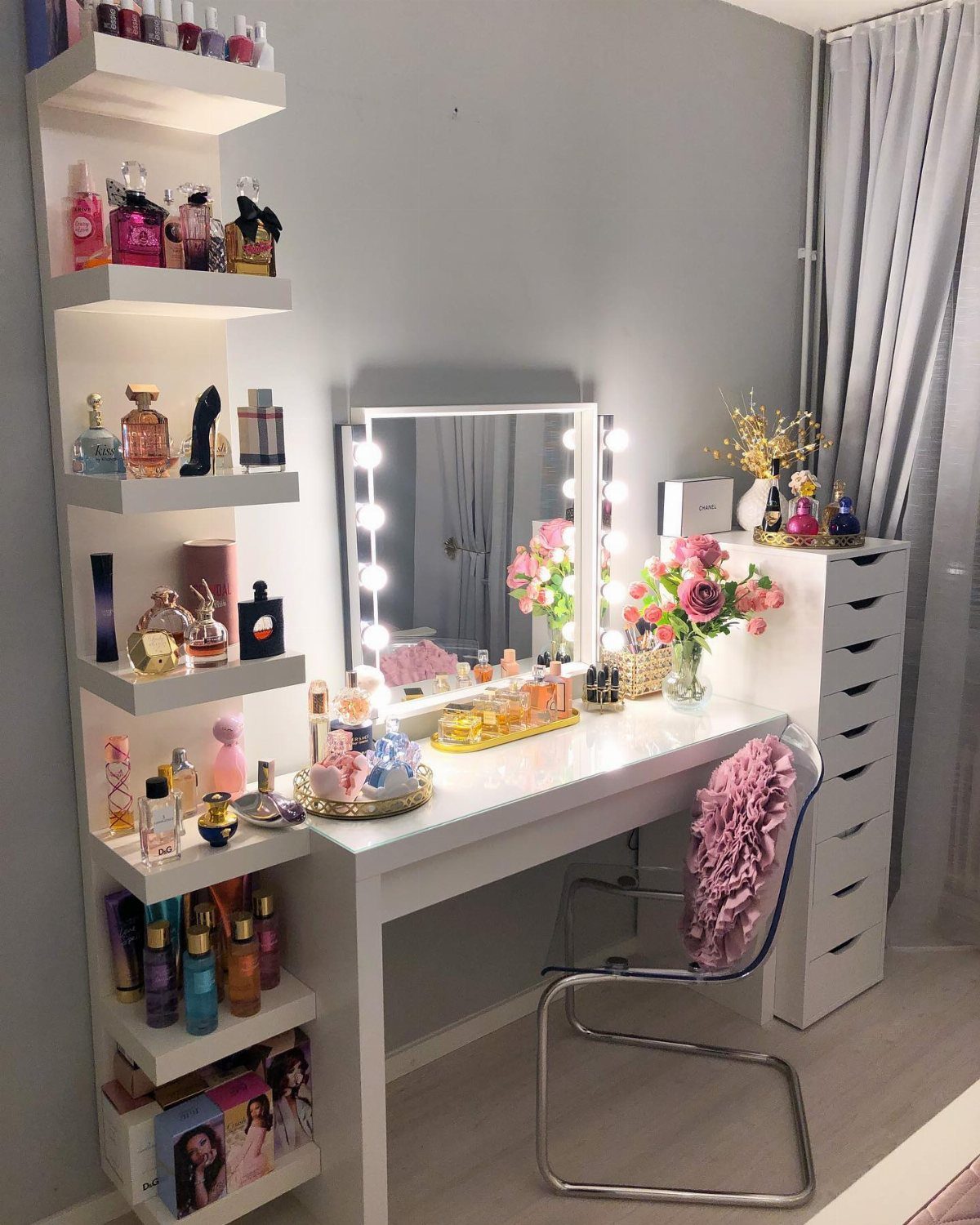 A white vanity desk with a lighted mirror, perfumes, and pink flowers in a modern room.
