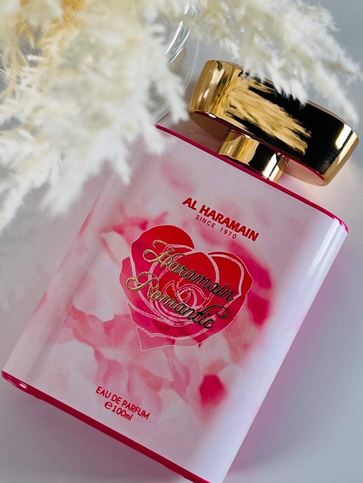 A pink Al Haramain Romantic perfume bottle with a gold cap near white pampas grass.
