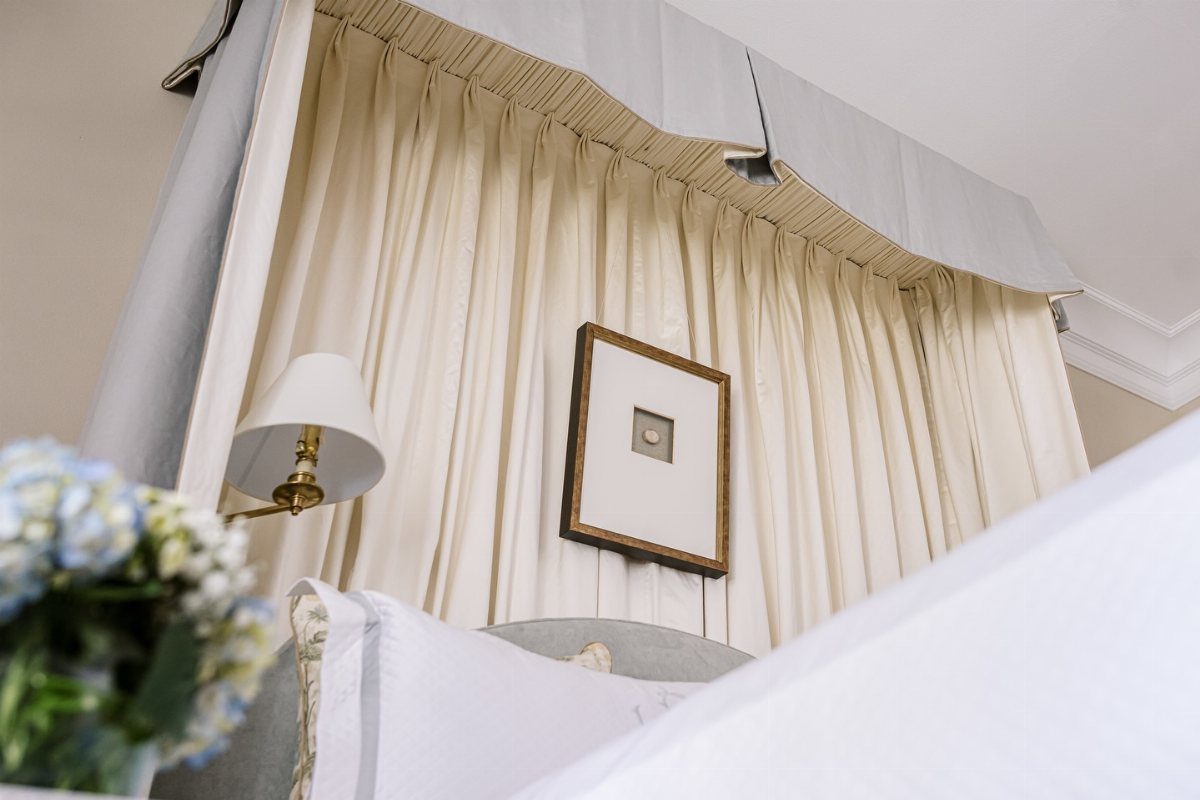 Cream fabric drapes hanging from a canopy bed frame against a neutral wall.