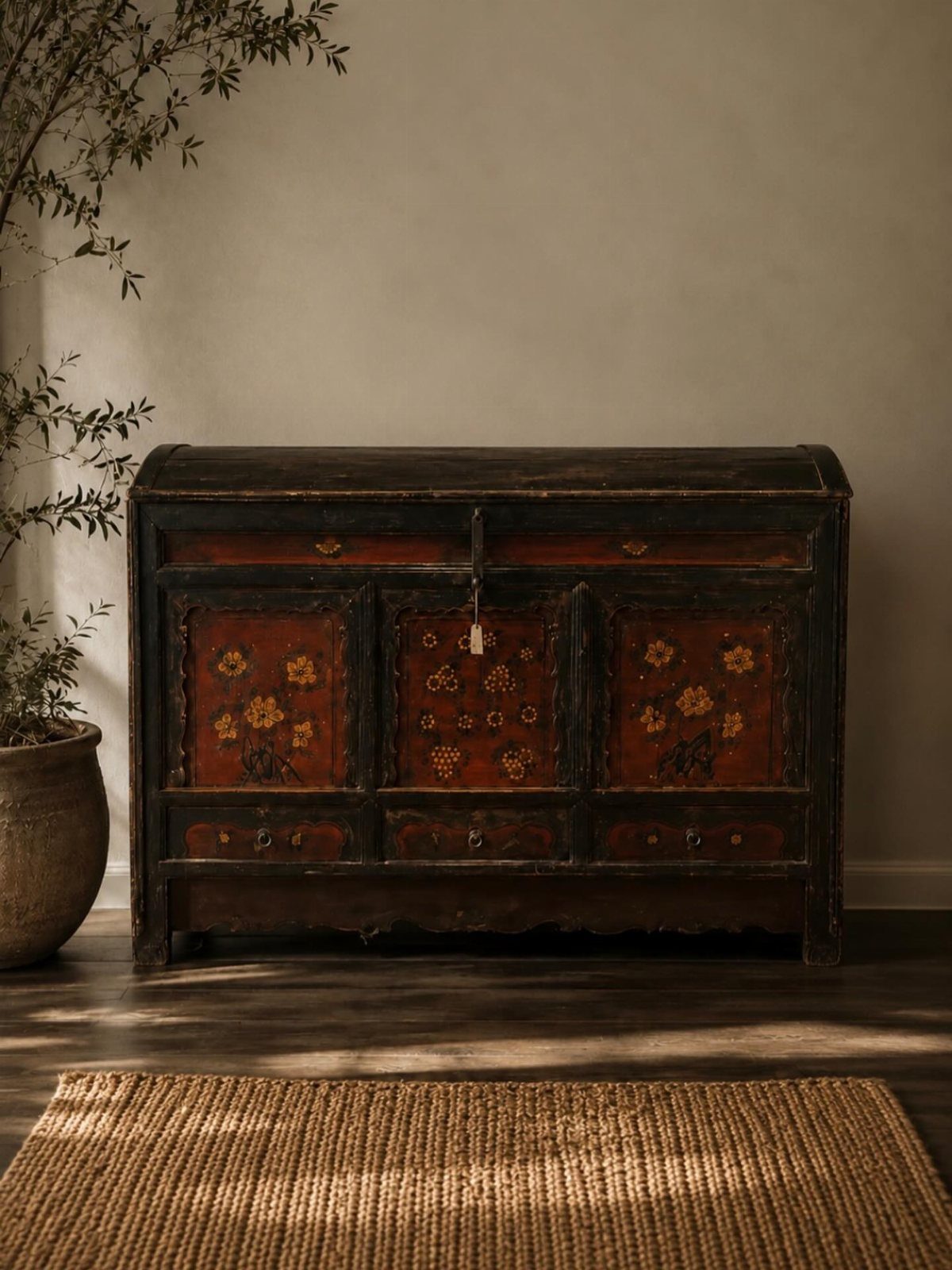 Ornate vintage wooden chest with red floral panels sitting on a dark floor near a plant.