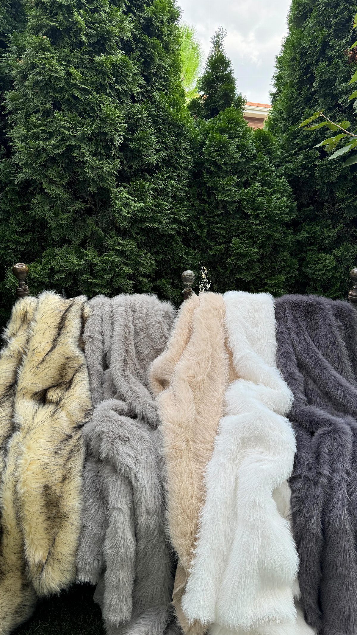Five textured faux fur throws in various neutral colors draped against a green hedge.