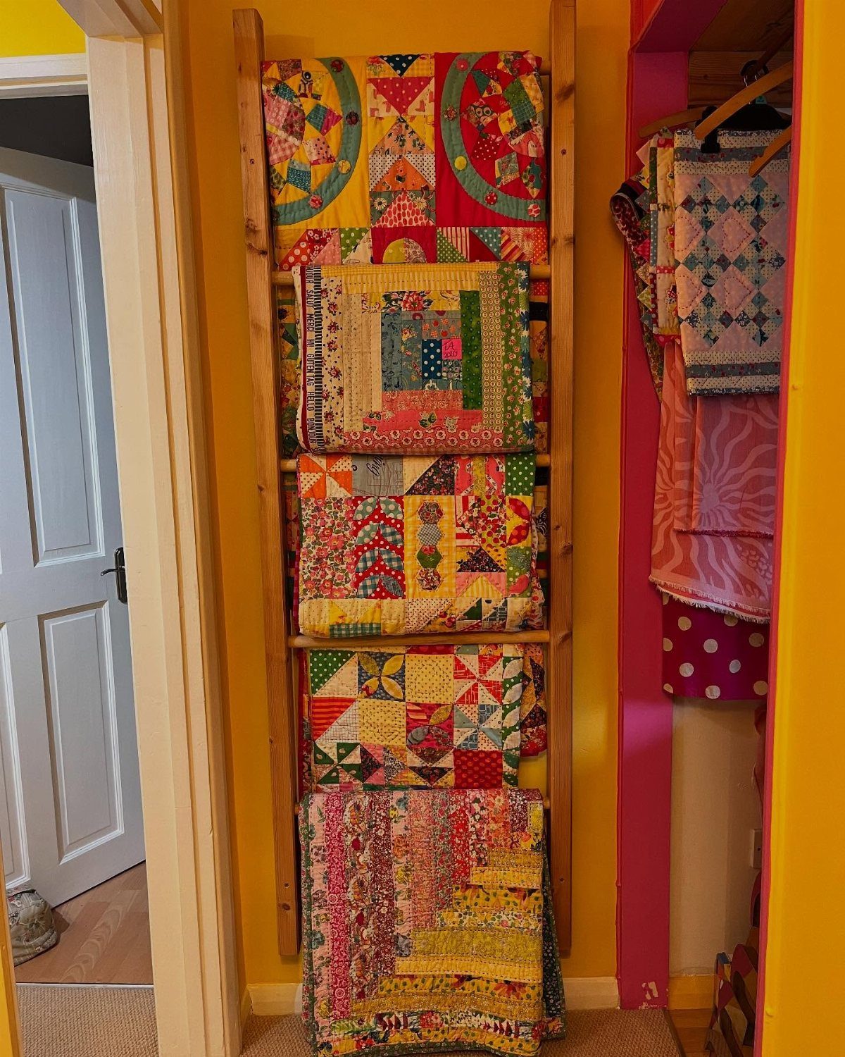 Colorful patchwork quilts hanging on a wooden ladder rack against a bright yellow wall.