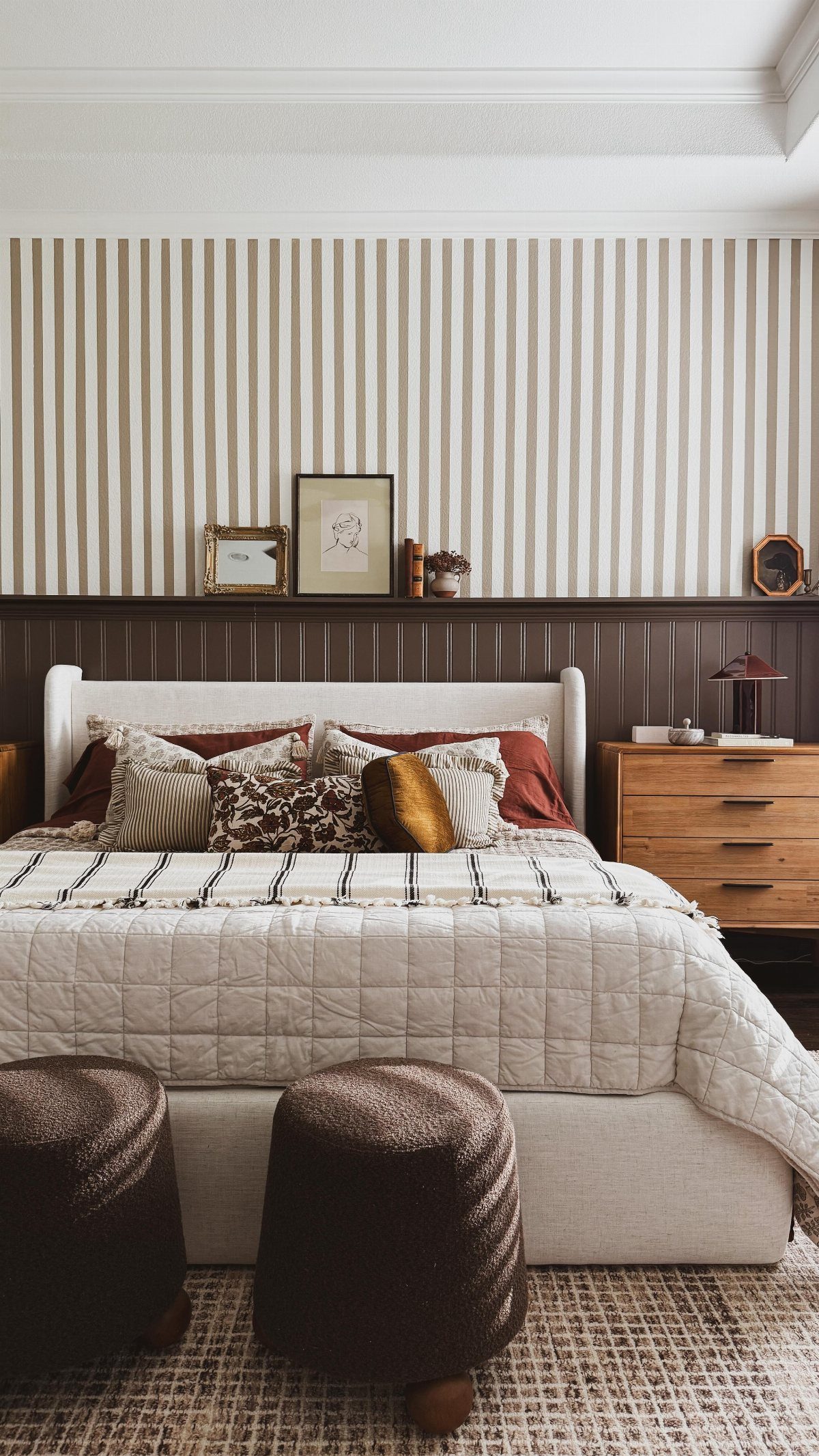Bed with patterned pillows set against dark brown beadboard paneling and striped wallpaper.