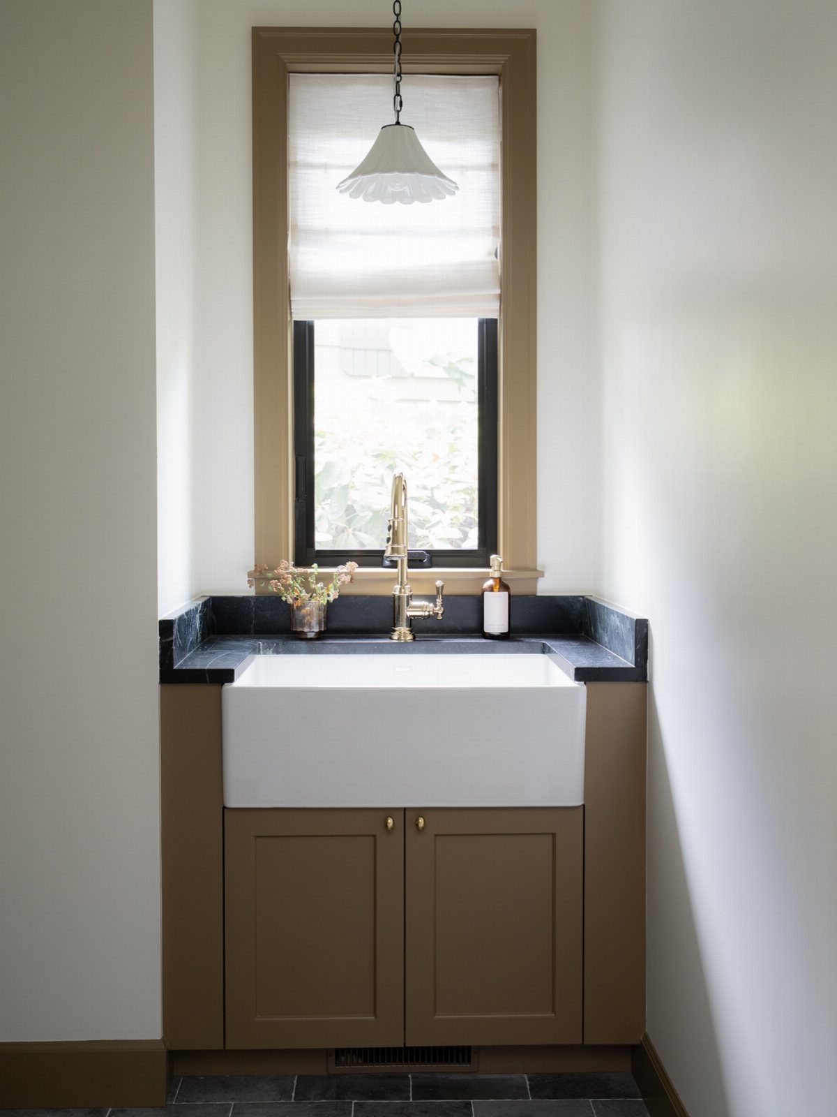 White ceramic farmhouse sink with a gold faucet set into a dark countertop near a window.