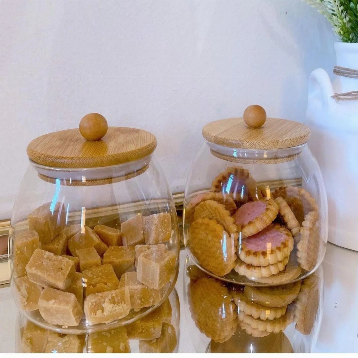 Two glass canisters with wooden lids containing fudge and cookies on a reflective surface.