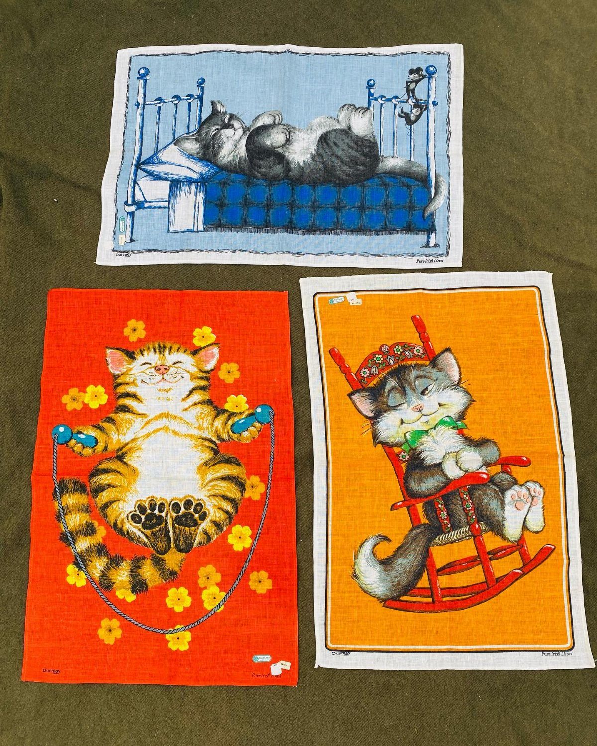 Three colorful cartoon cat dish towels laid flat on a dark green fabric background.