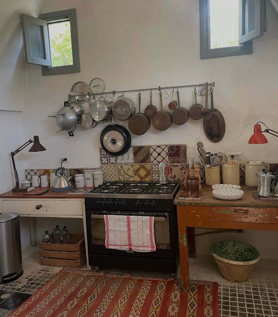 Metal pot rack with copper and silver cookware hanging above a rustic kitchen stove.