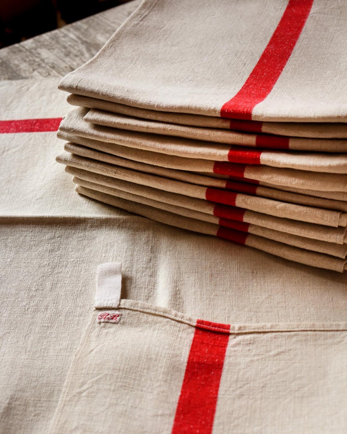 Stacked beige linen dish towels with a bold red stripe on a textured surface.