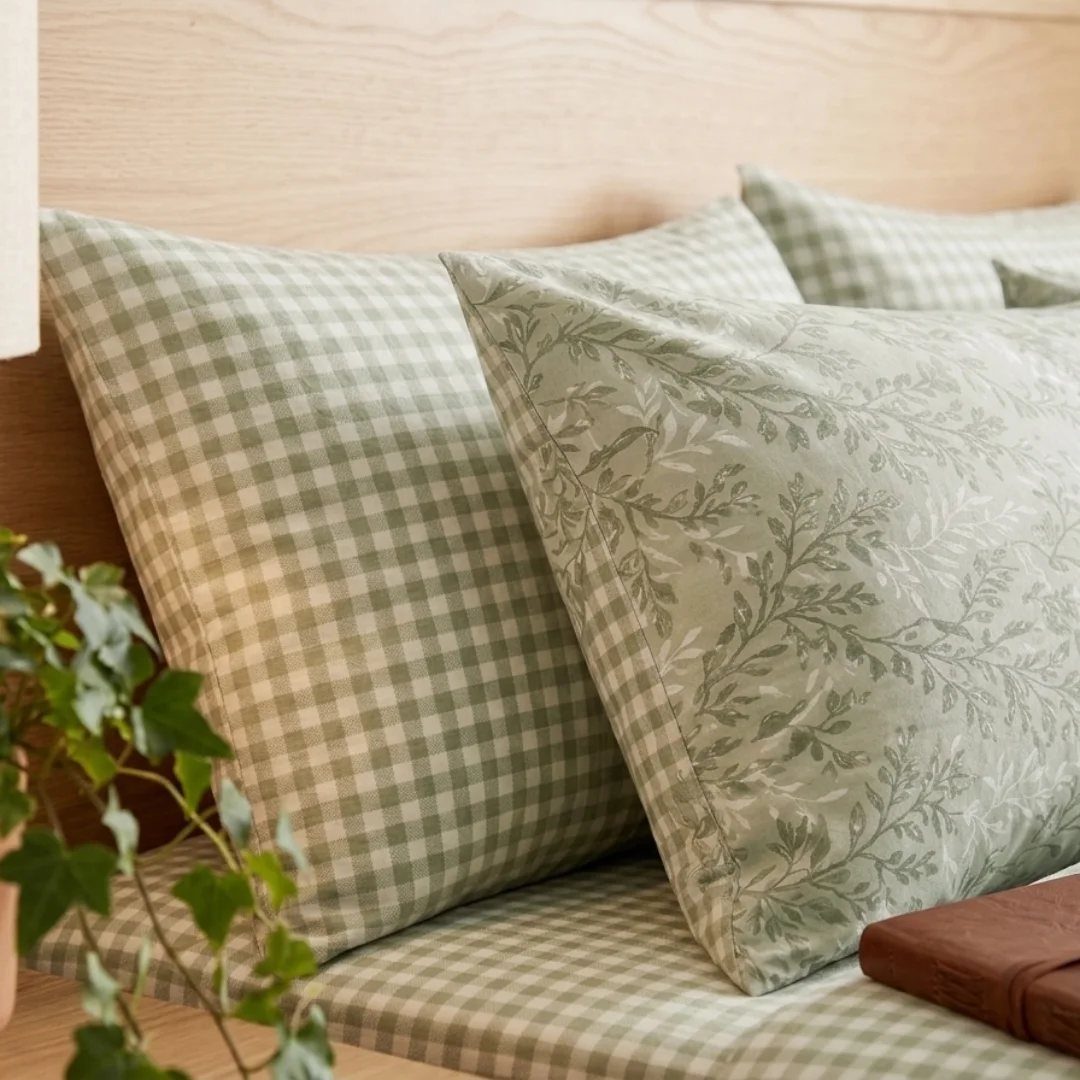 Sage green gingham and botanical patterned pillows resting on a bed with a wooden headboard.