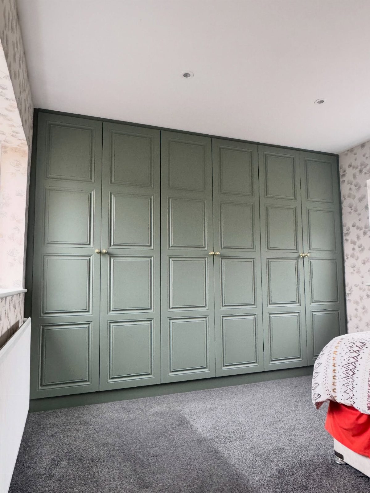 Sage green paneled built-in wardrobes with gold knobs against patterned wallpaper.