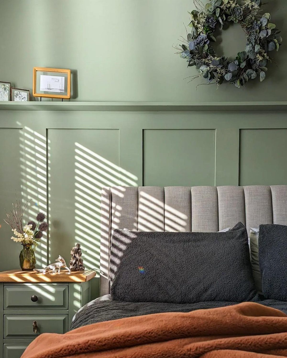 Sage green paneled walls behind a gray upholstered headboard and dark pillows in a bedroom.