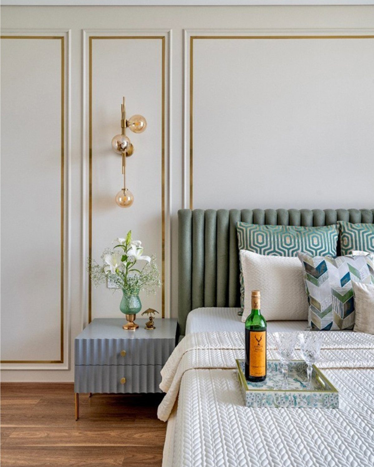Sage green upholstered headboard and patterned pillows next to a gold wall sconce with warm lighting.