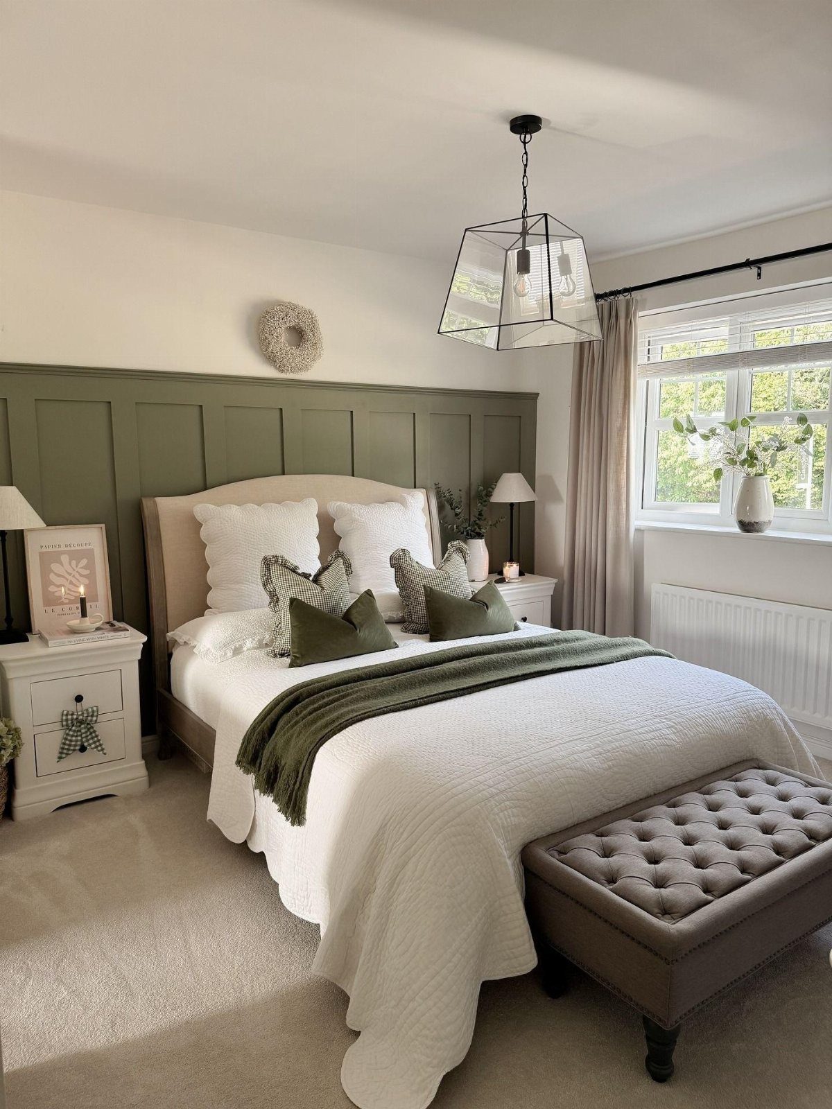 Sage green paneled walls behind a white bed with green accents and a grey tufted ottoman.