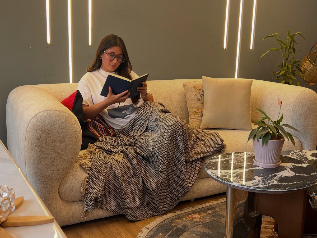 A woman wrapped in a textured blanket reads a book while sitting on a cozy cream sofa.