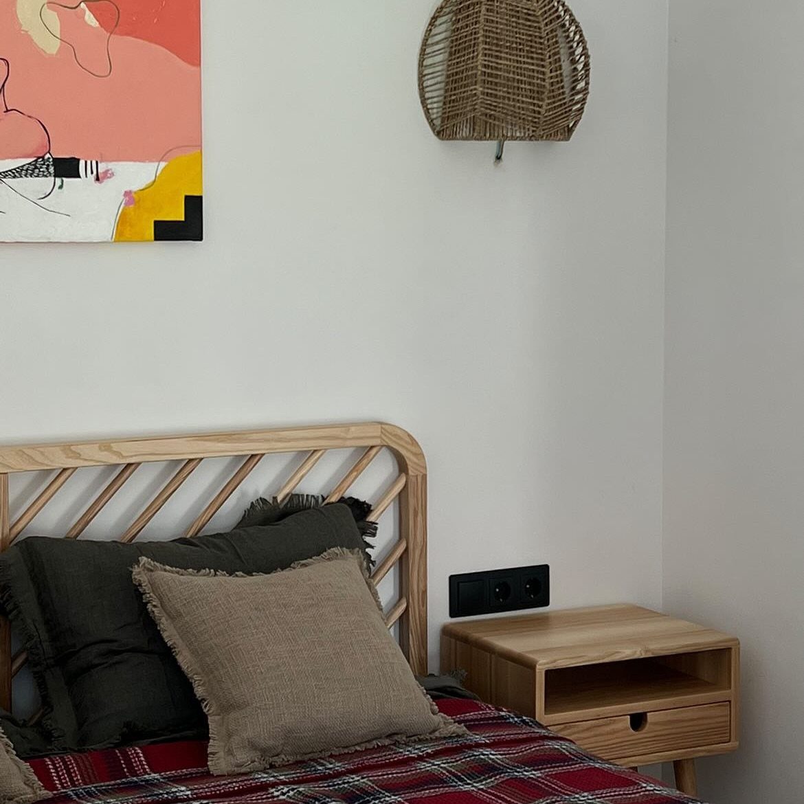 Light wood bed frame with textured pillows and a plaid blanket against a white wall.