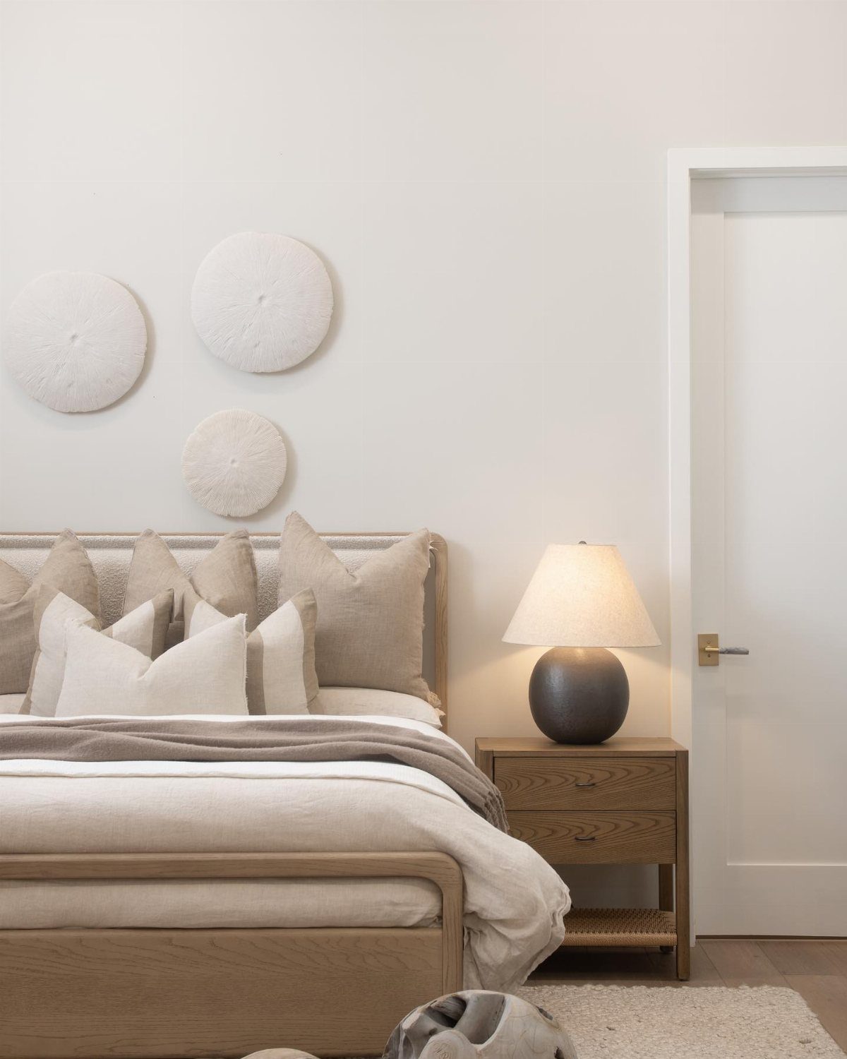 Minimalist bedroom with light wood bed, neutral linen bedding, and circular textured wall art.