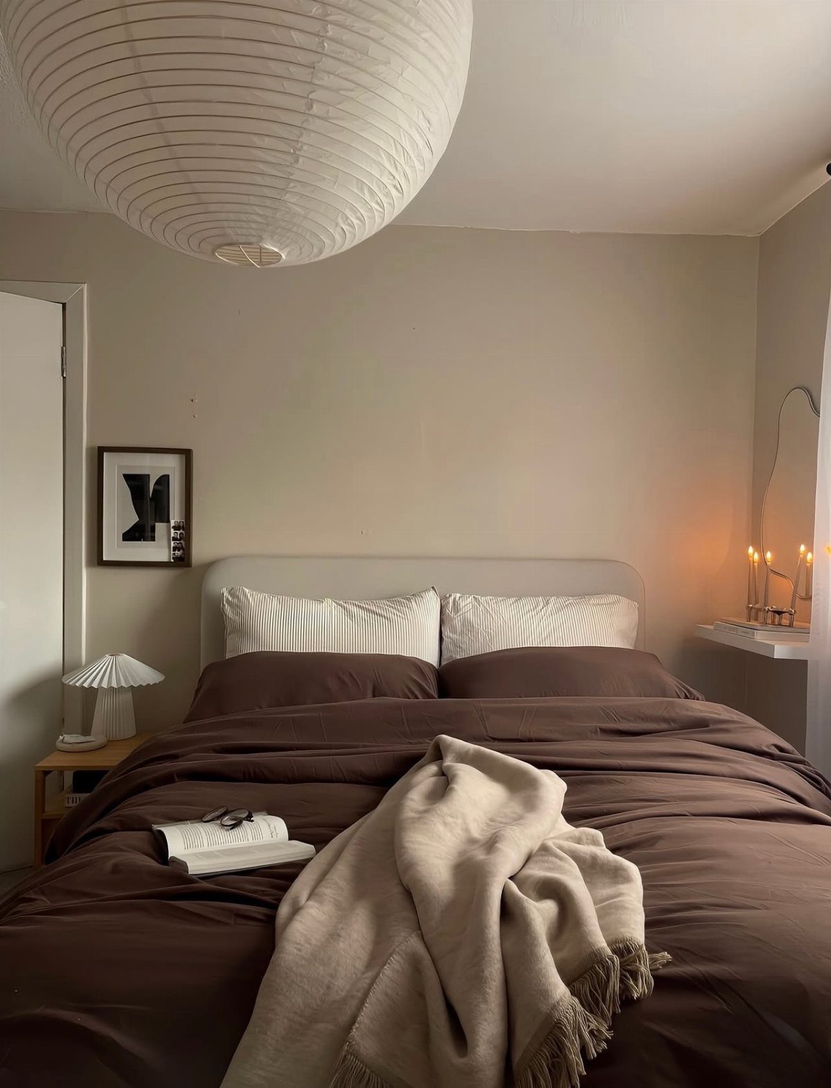 A cozy bedroom with dark brown bedding, white pillows, and a large white paper lantern hanging above.