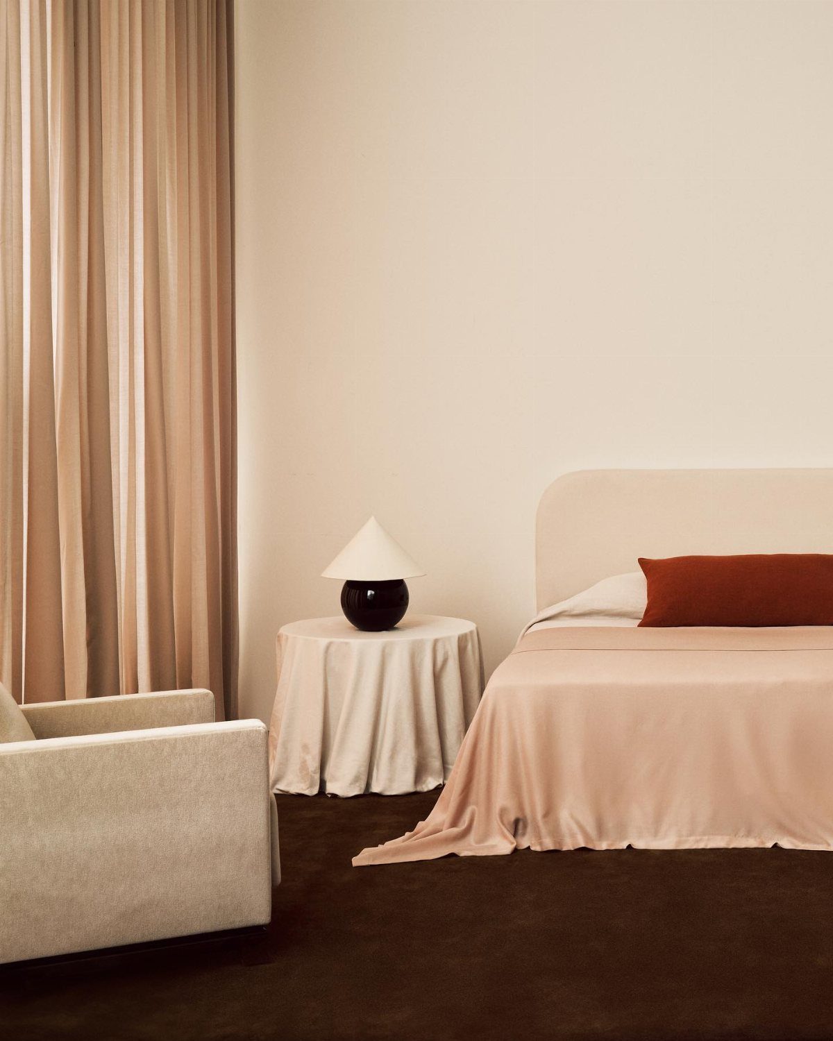 Minimalist bedroom with peach bedding, a small side table with a lamp, and neutral curtains.