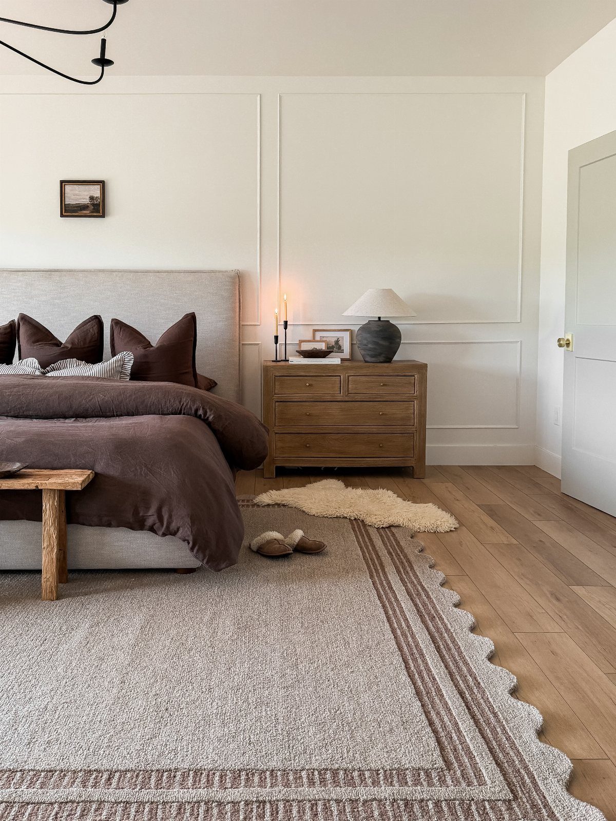 A cozy bedroom with dark brown bedding, a wooden nightstand, and a large textured rug on wood floors.