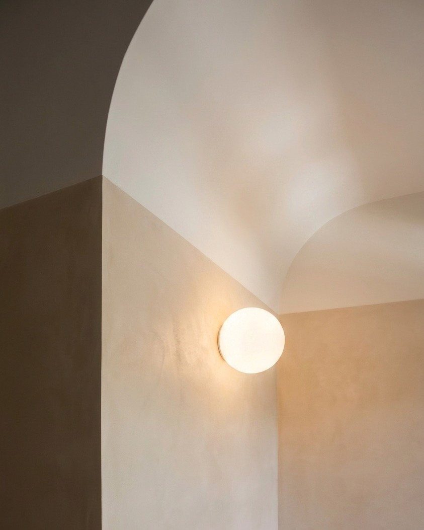 A glowing white spherical wall sconce illuminating a minimalist beige arched wall.