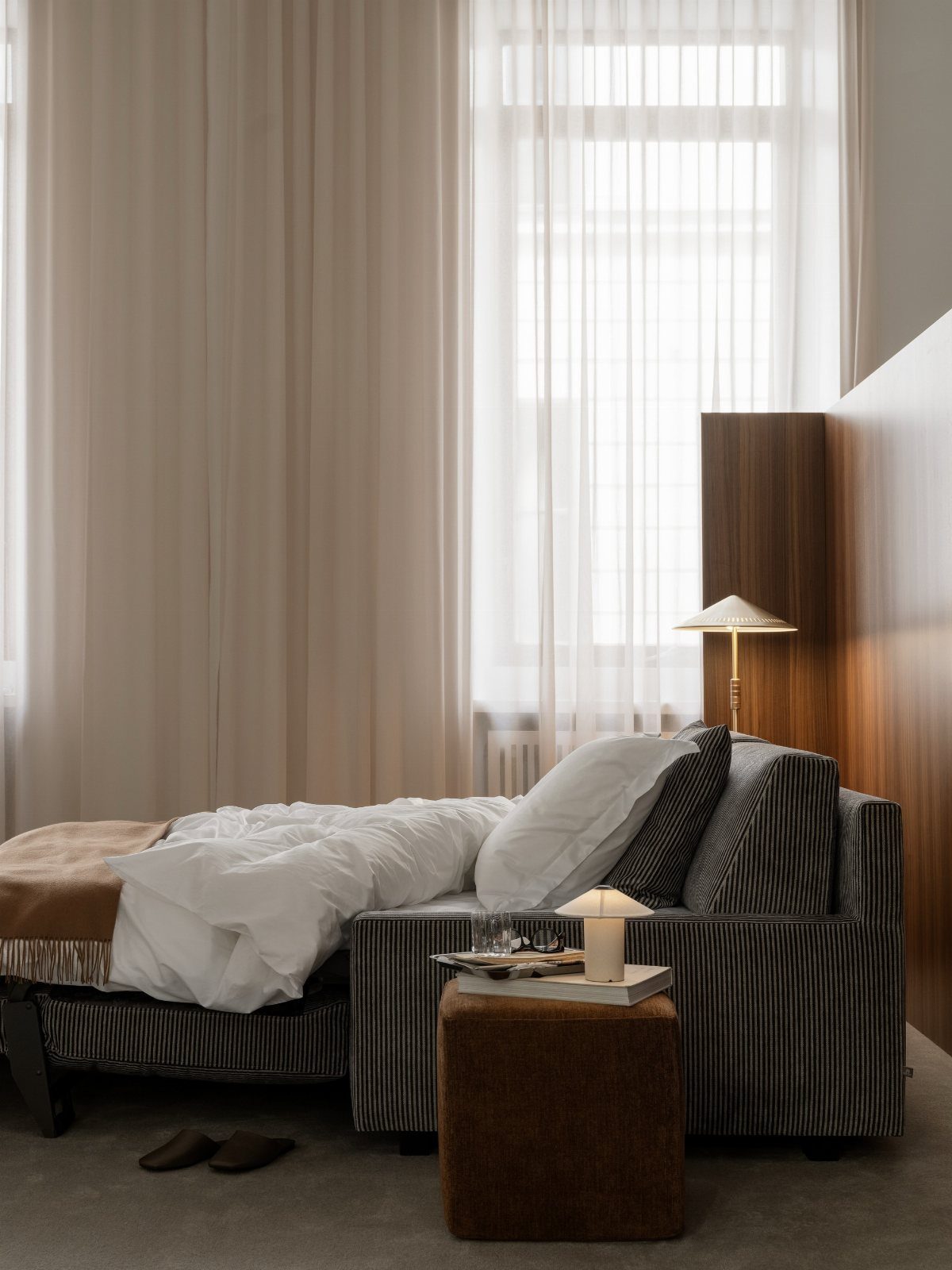 A low bed with striped upholstery and white linen bedding sits near sheer curtains.