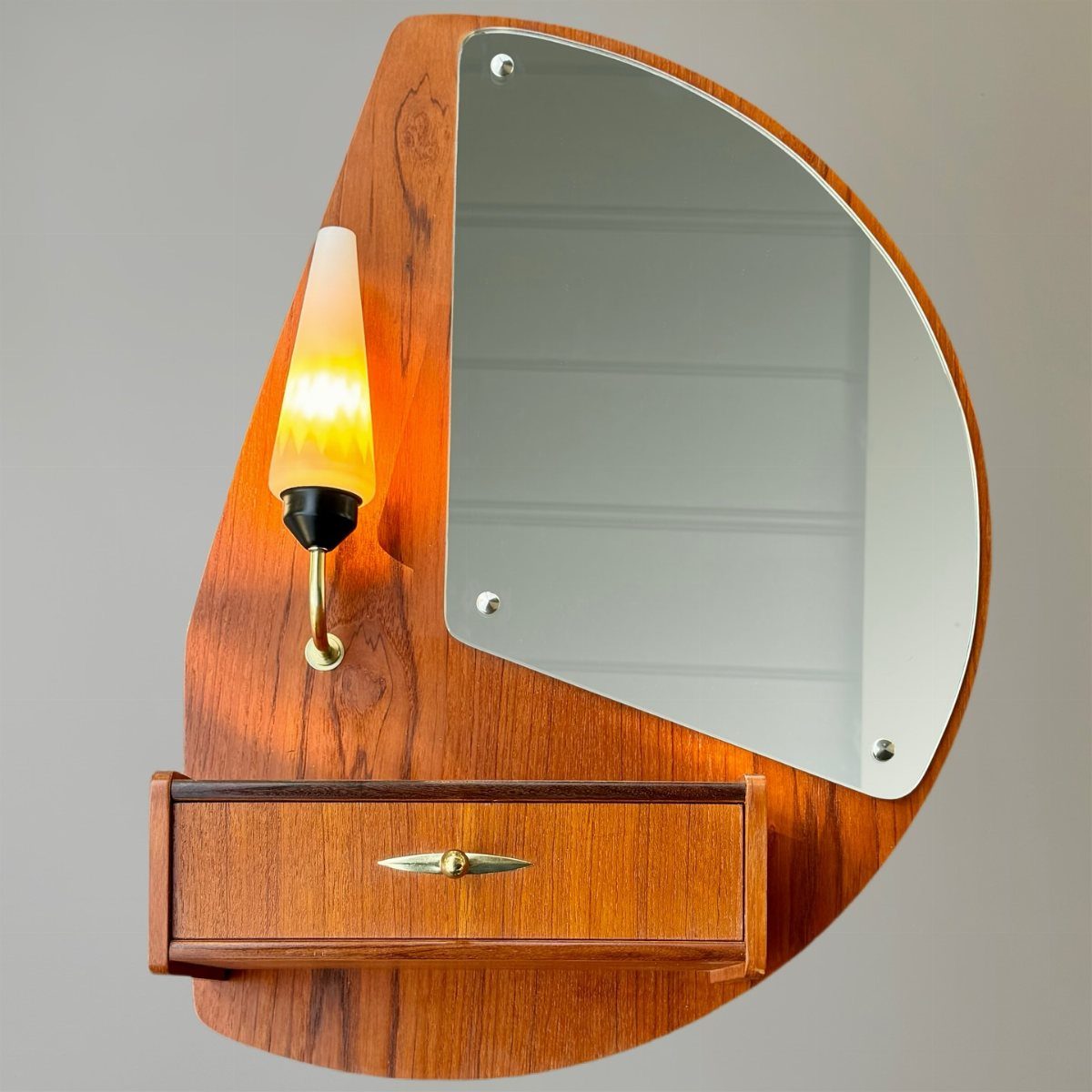 Mid-century modern wooden wall unit with an asymmetrical mirror, a small drawer, and a glowing amber lamp.