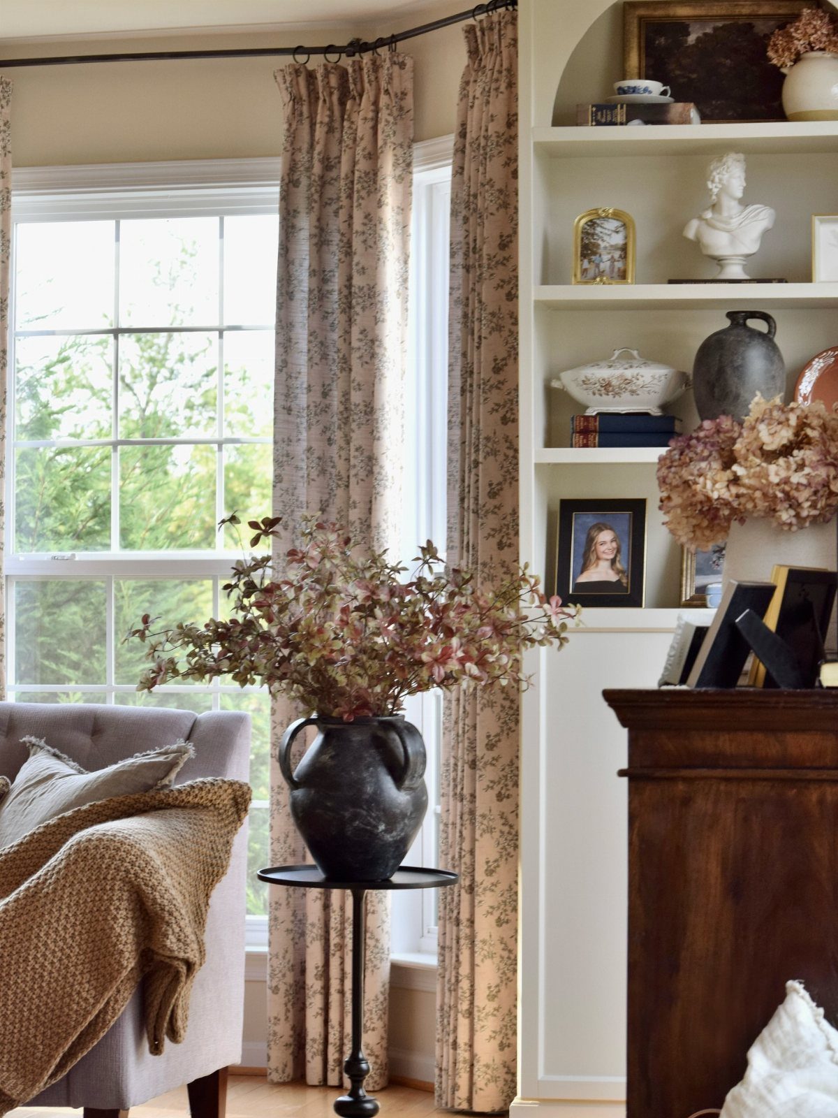 Floral patterned curtains hang beside a window near a grey armchair with a textured throw.