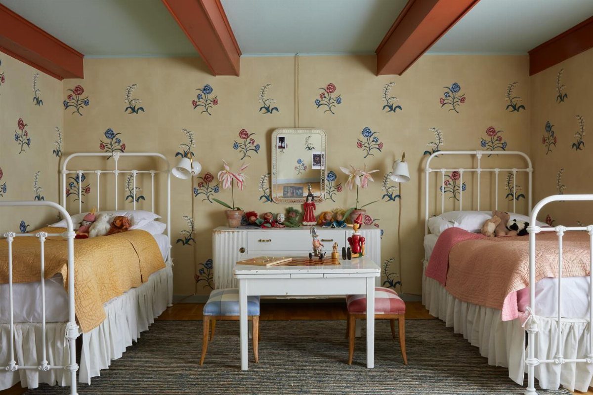 Two white metal beds with colorful linens in a room with floral wallpaper and red beams.