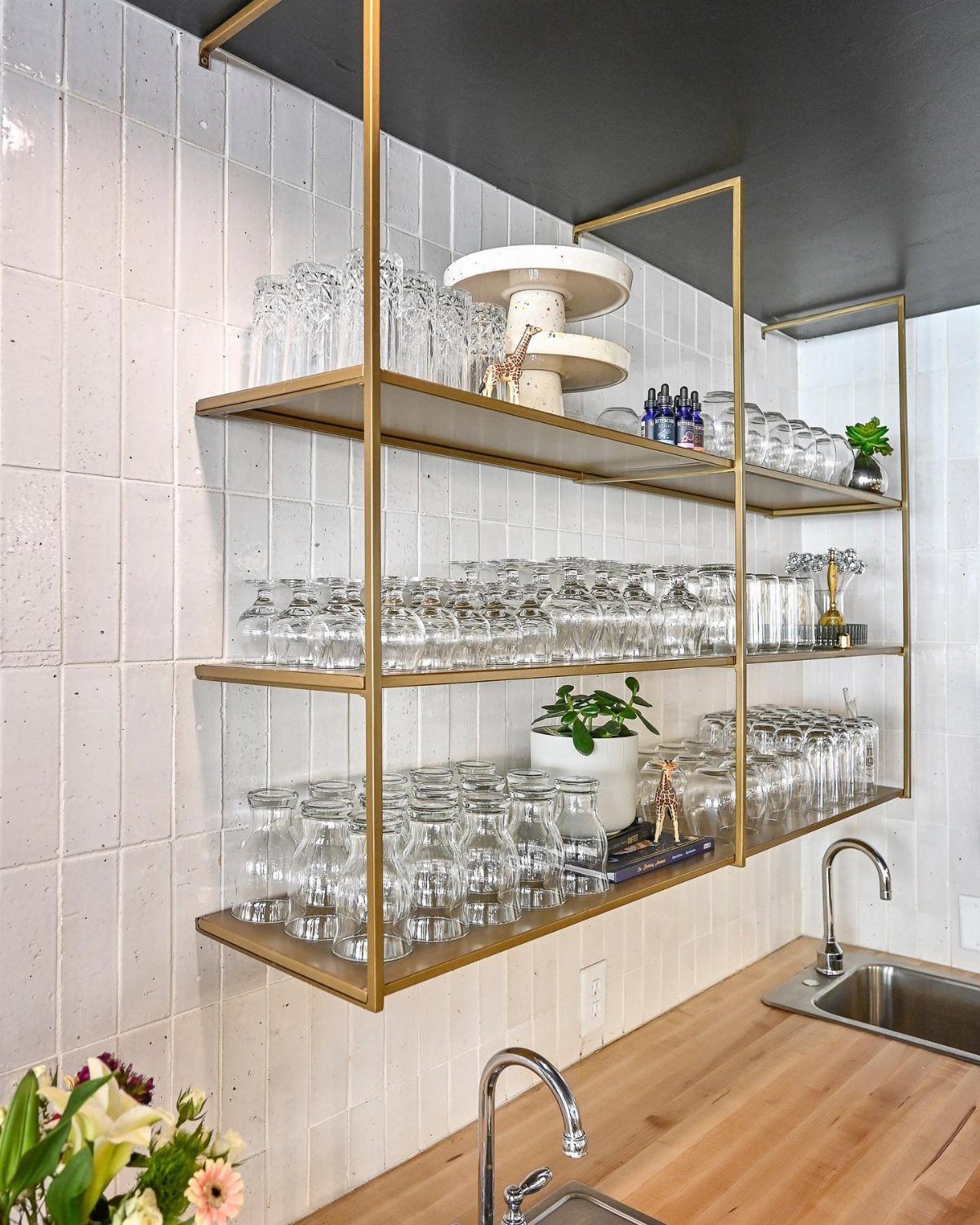 Gold metal shelving units holding various clear glassware and plants against a white tiled wall.
