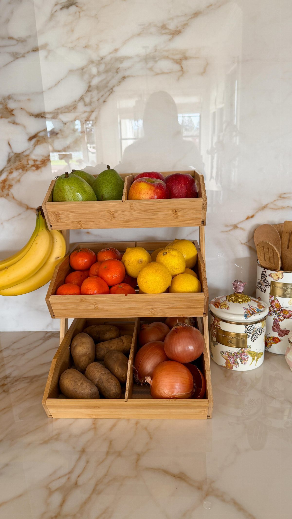 Three-tiered bamboo organizer filled with fresh fruits and vegetables on a marble countertop.