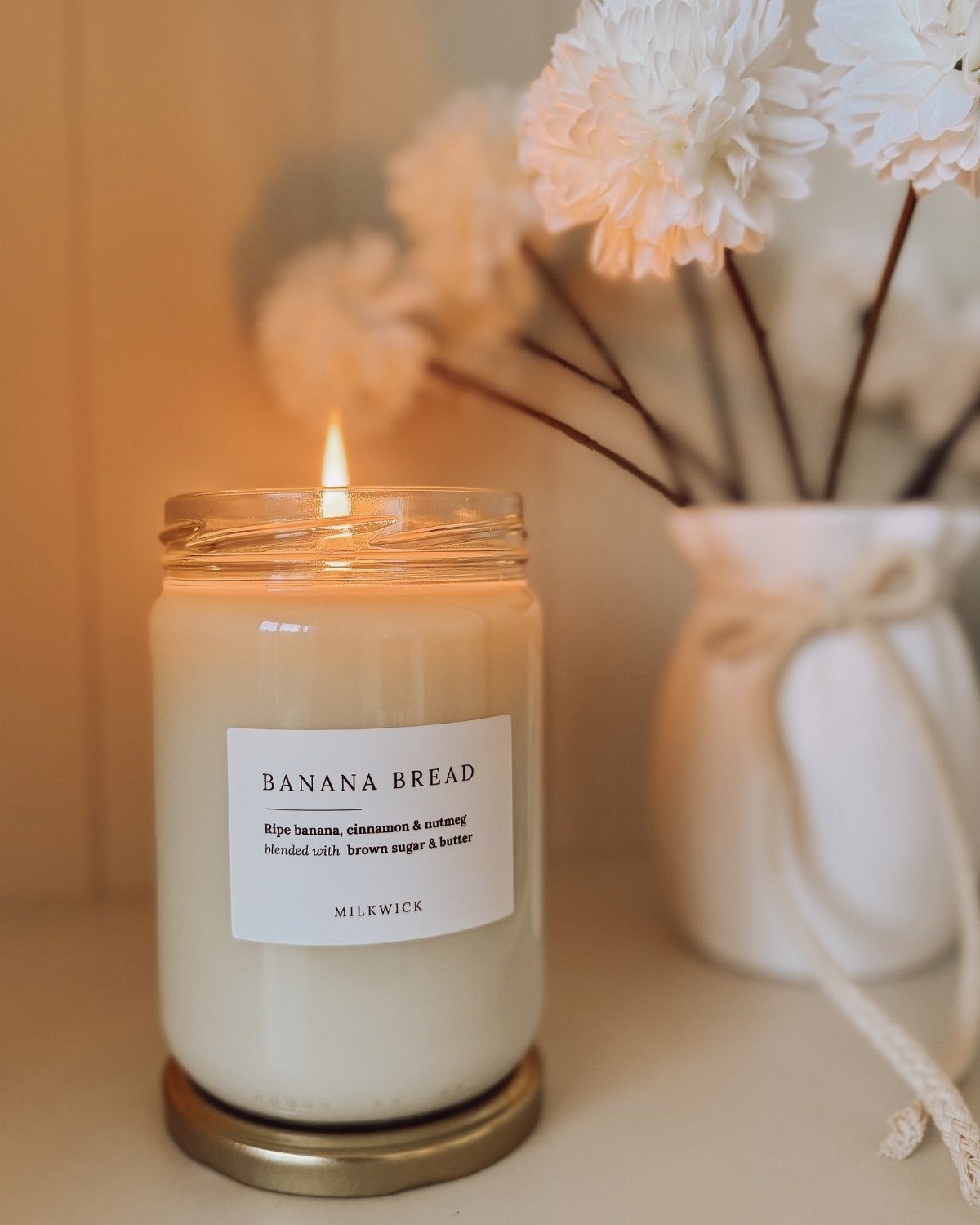 A lit banana bread scented candle in a glass jar next to white flowers.