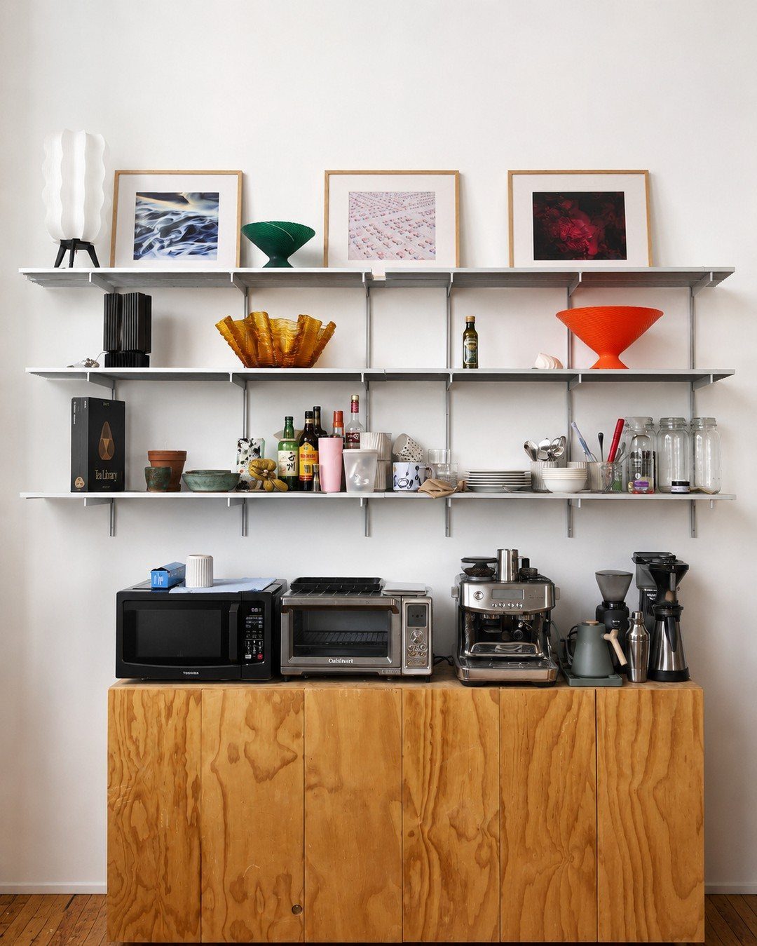 White metal wall shelves holding colorful bowls, framed art, and various kitchen items above a wooden sideboard.