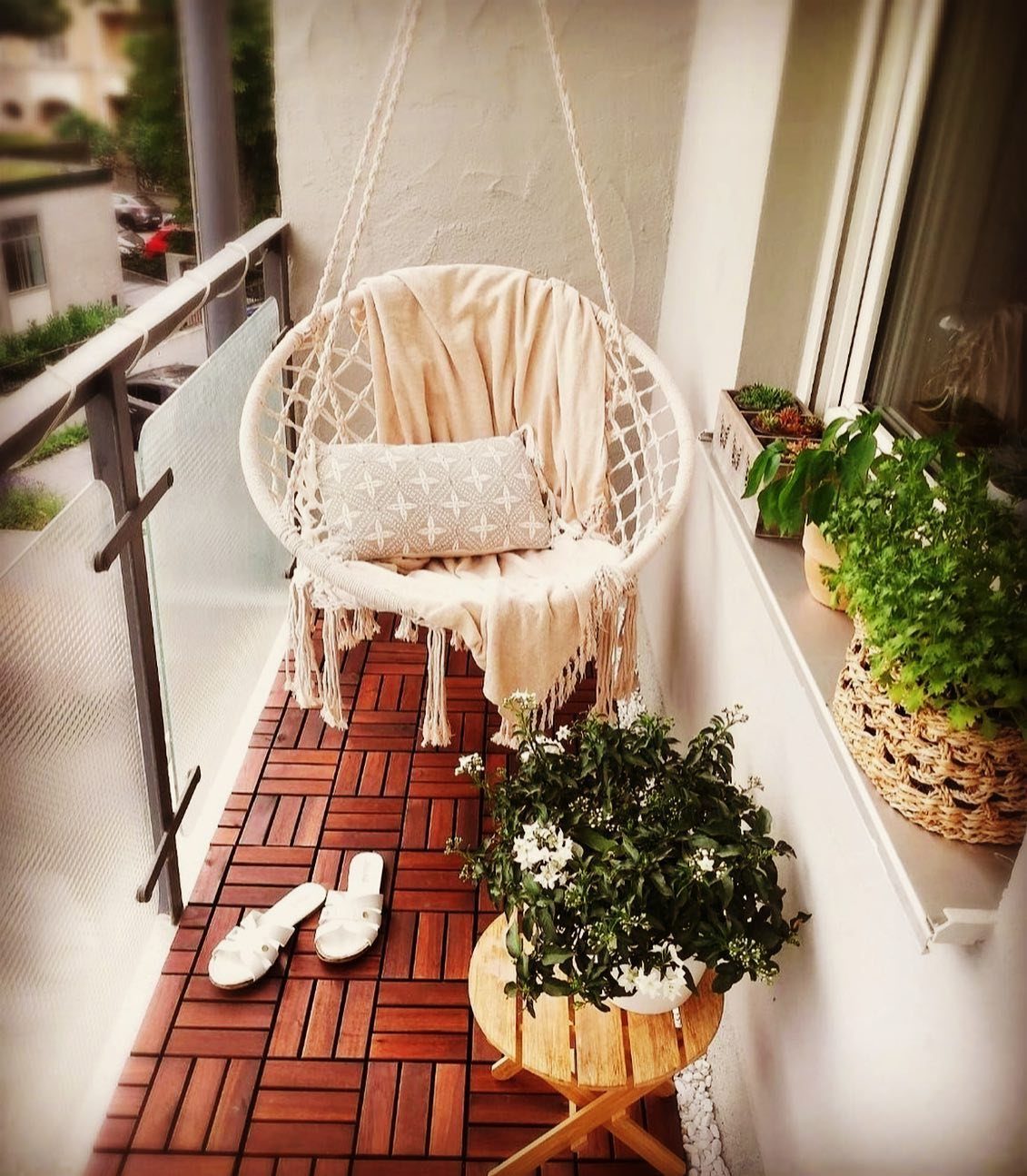 A white macramé hanging egg chair with cushions on a small decorated balcony.