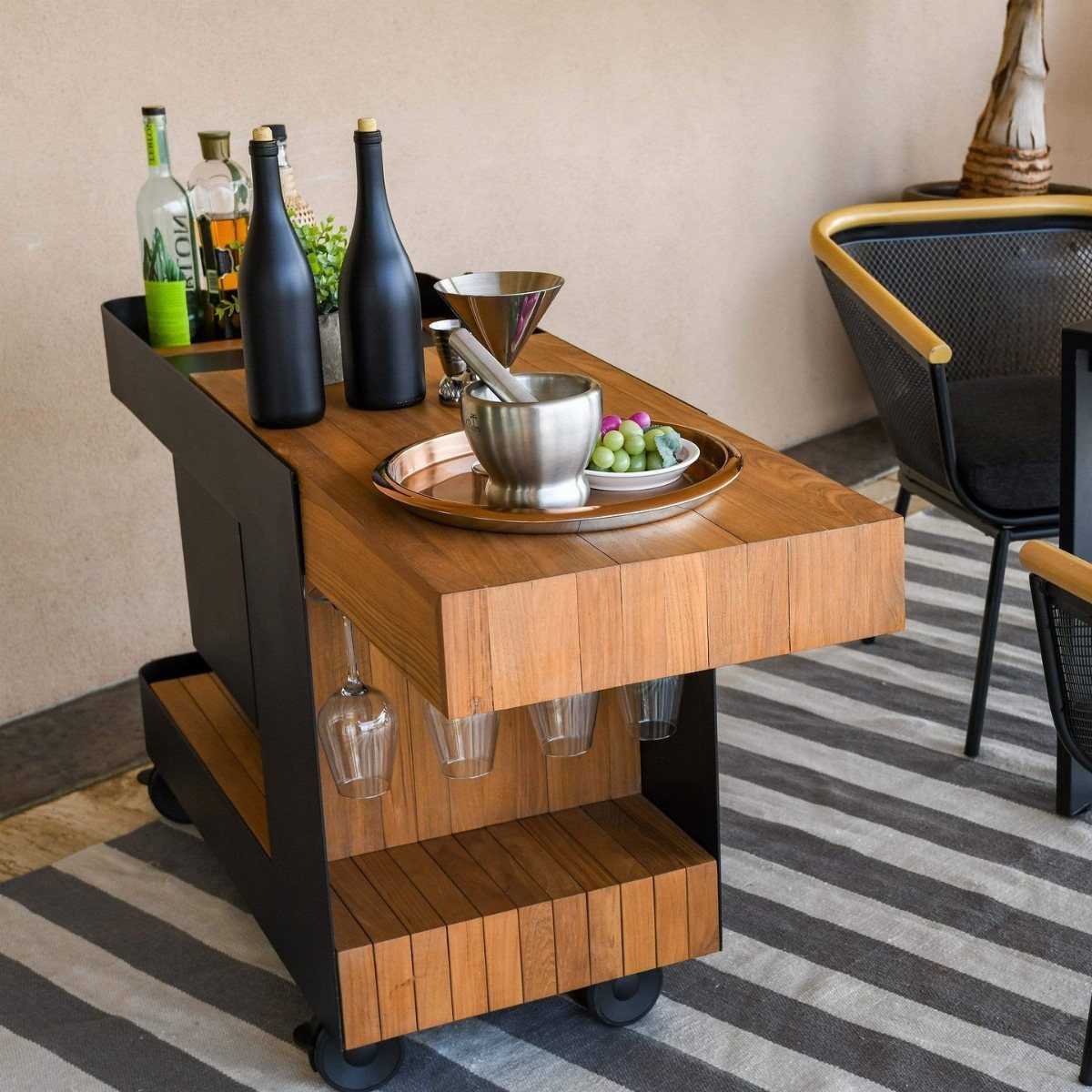 A wooden mobile bar cart stocked with wine bottles and silver serving ware on a rug.