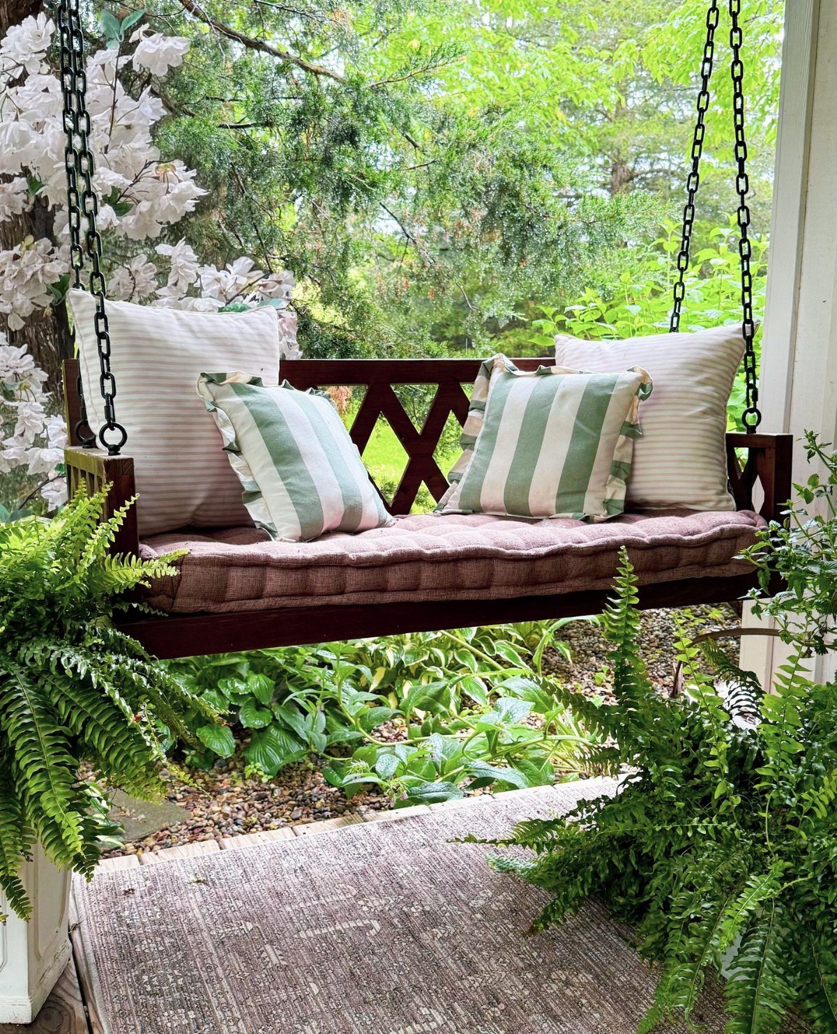 Wooden porch swing with striped pillows and a large cushion surrounded by green plants.