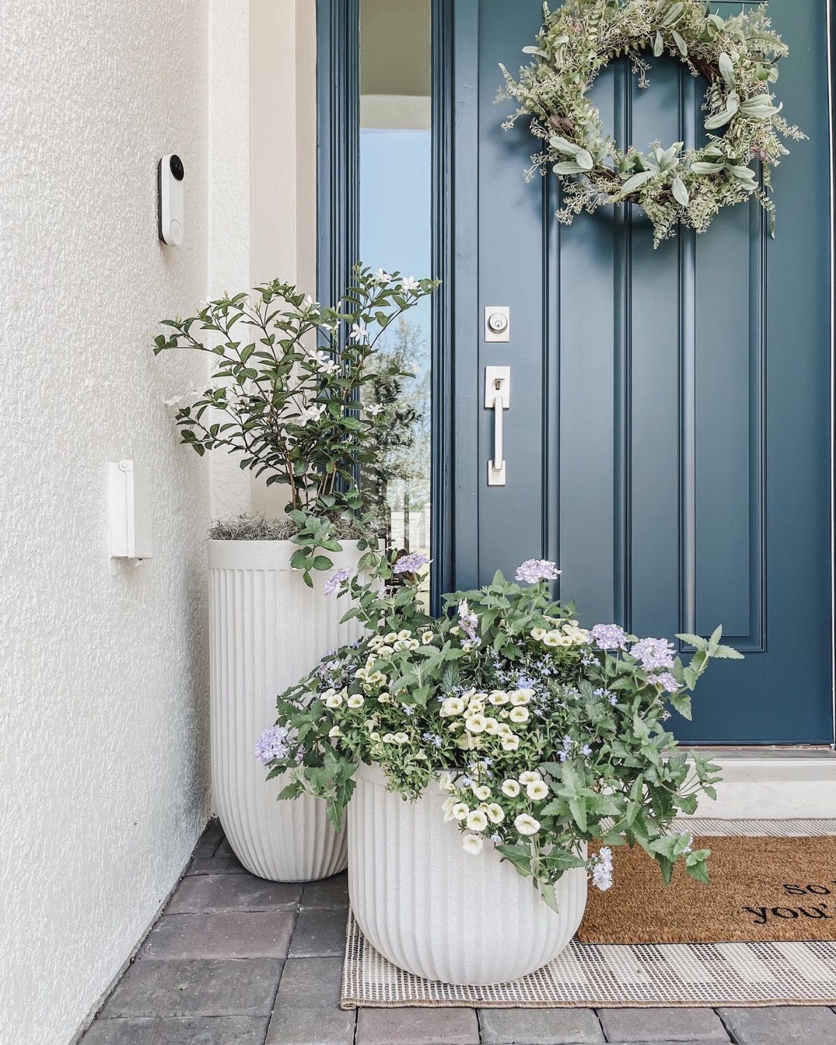White ribbed planters with flowers and a wreath on a blue door.