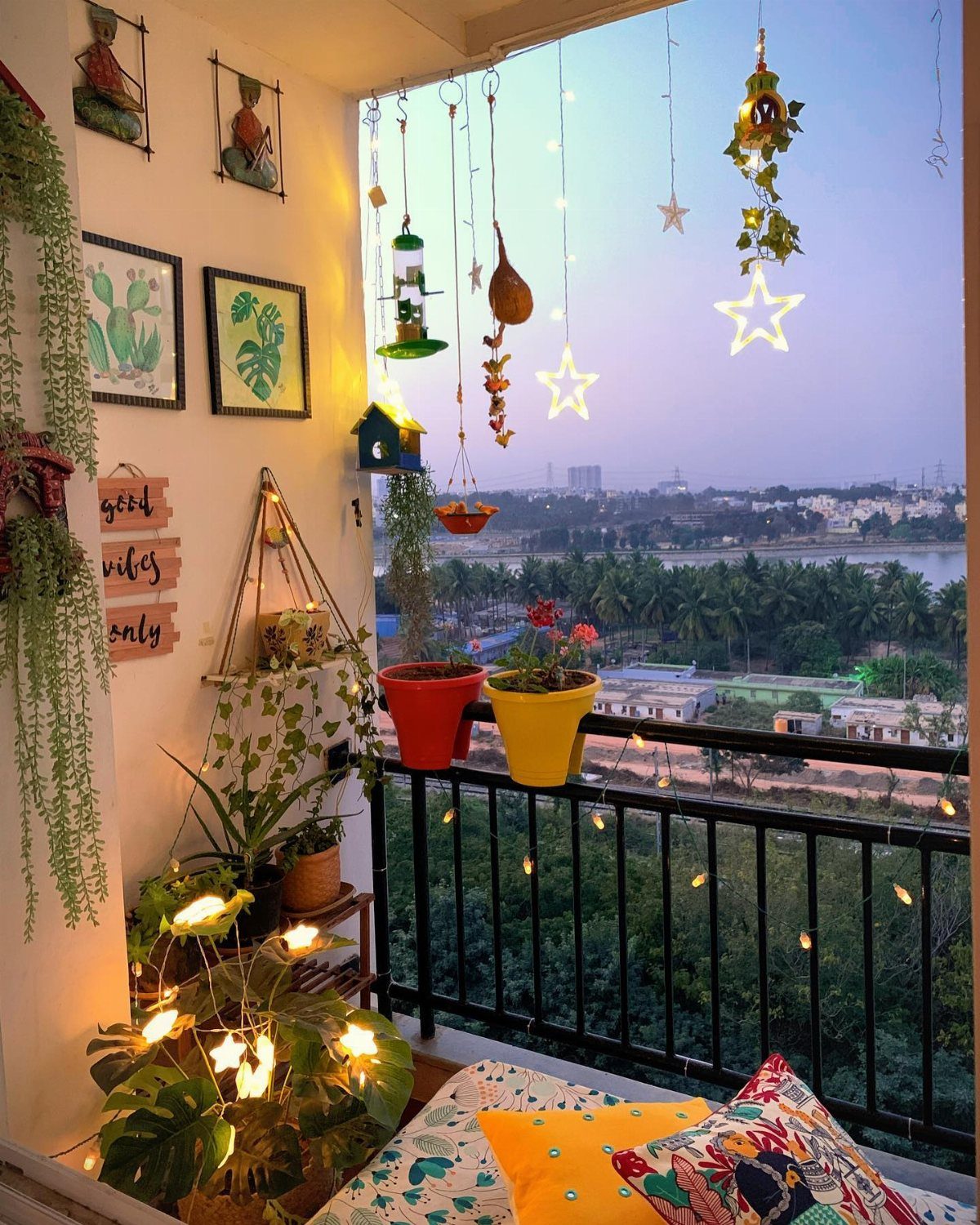 Small balcony decorated with hanging star lights, colorful potted plants, and patterned cushions at dusk.