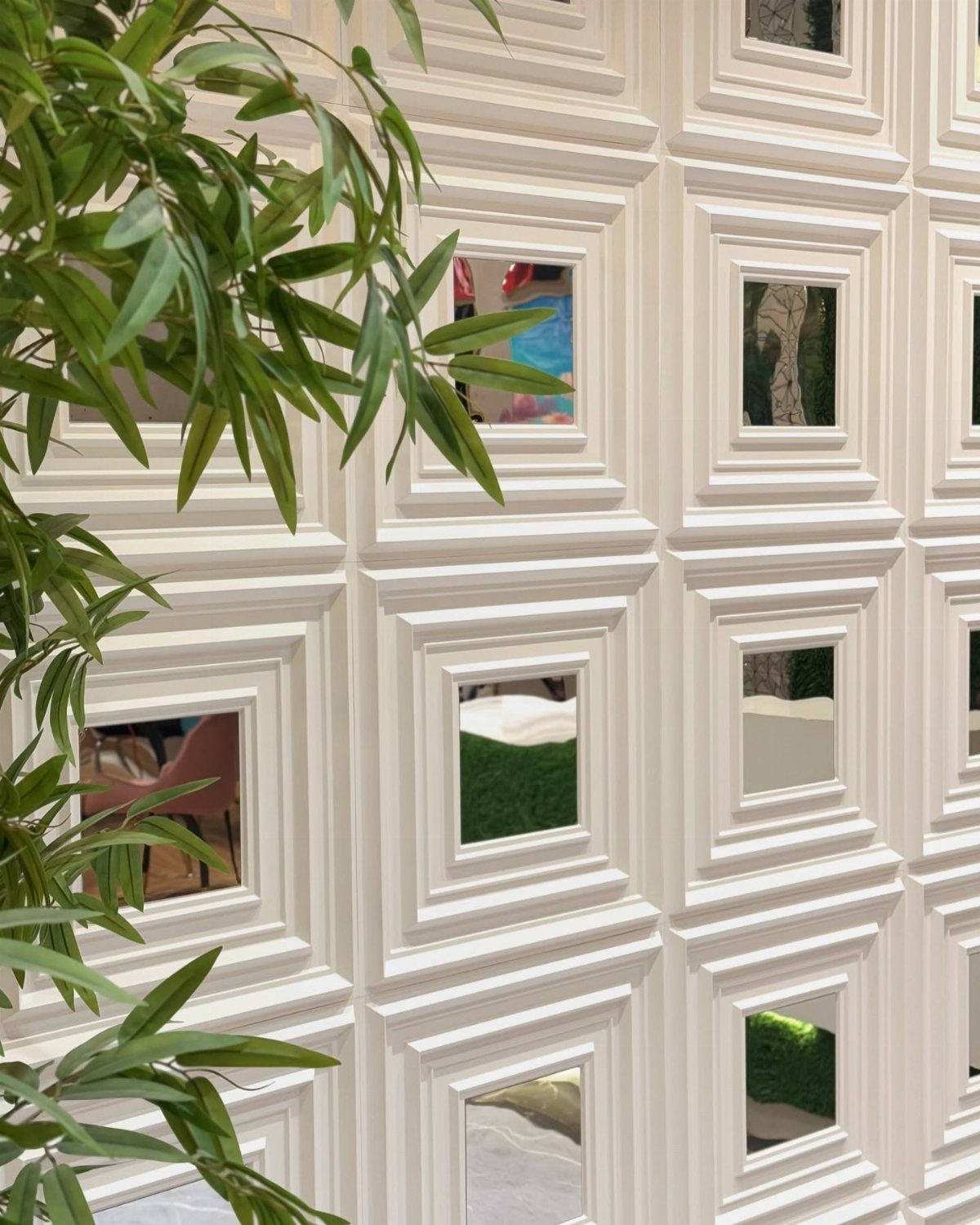 White paneled wall with small square mirrors and green leaves in the foreground.