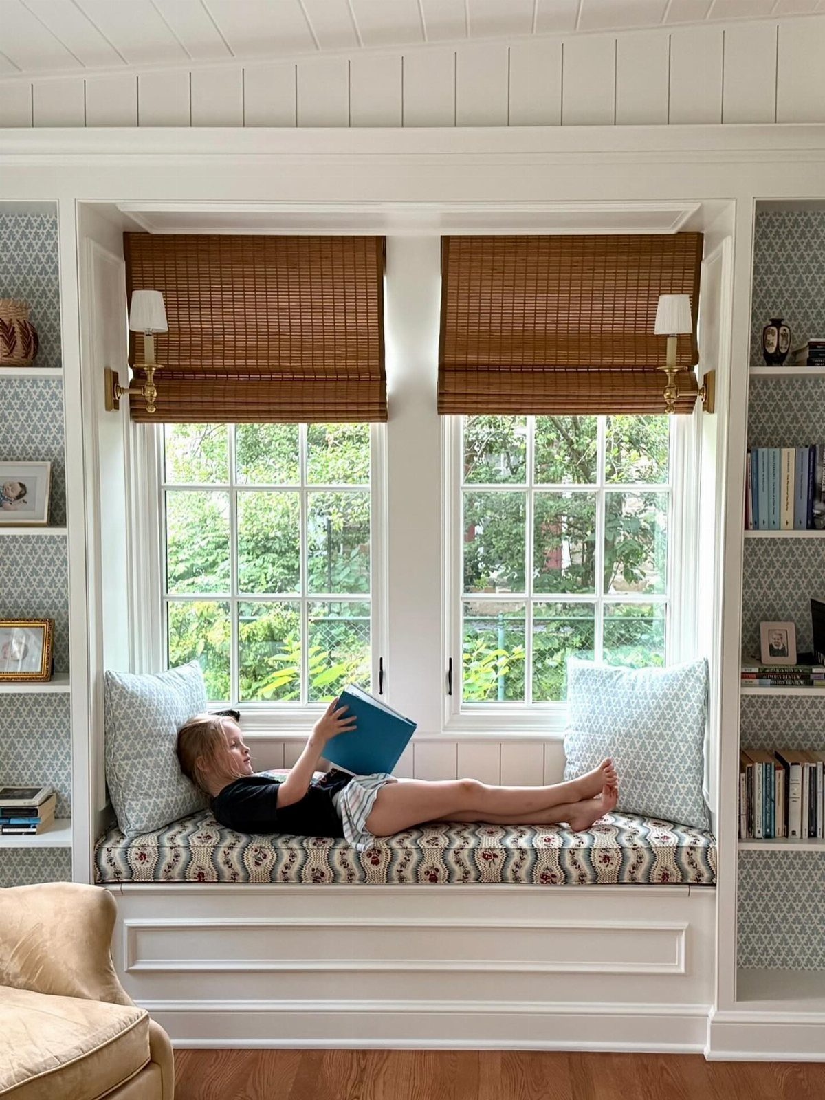 A young girl reading a blue book while lying on a built-in window seat.