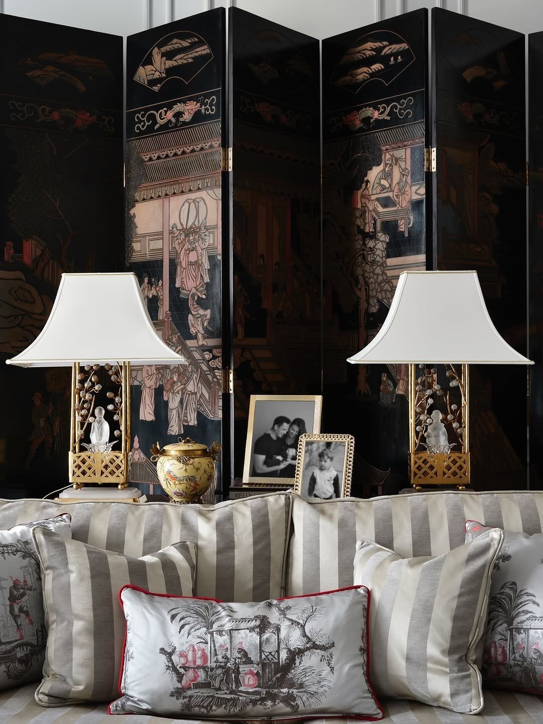 Black folding screen with traditional illustrations behind a table with lamps and framed photos.