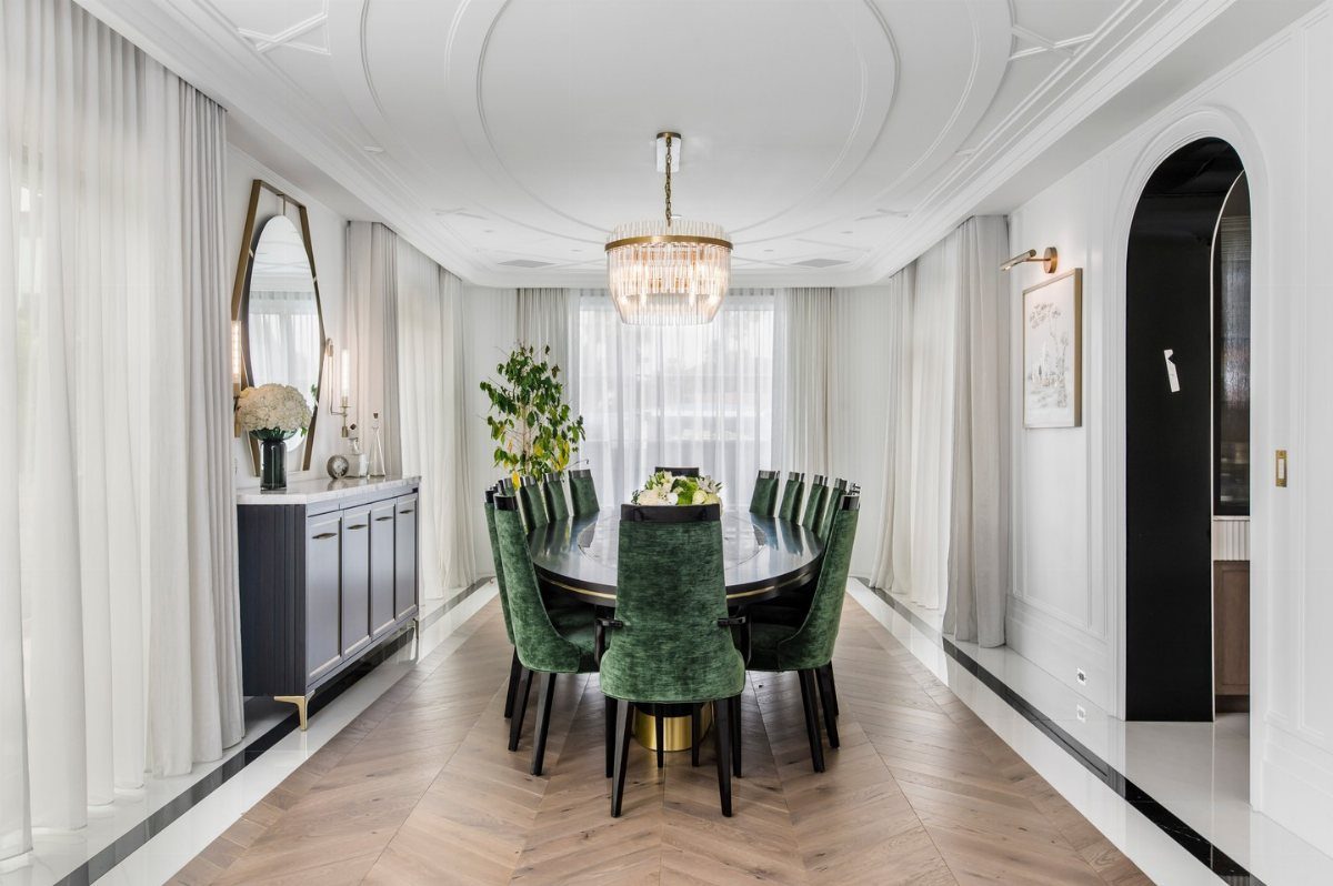 Green velvet dining chairs surrounding a large oval table in a bright, sunlit room.