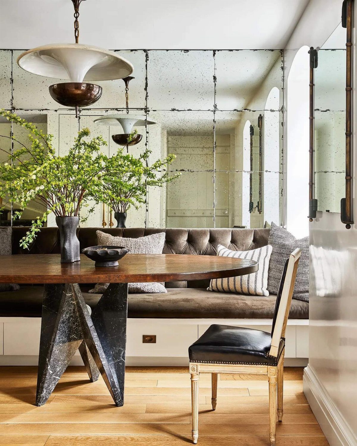 Round wooden dining table set against a large antiqued paneled mirror and tufted banquet seating.