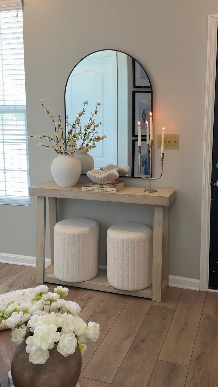 An arched mirror hangs above a light wood console table decorated with white vases and candles.