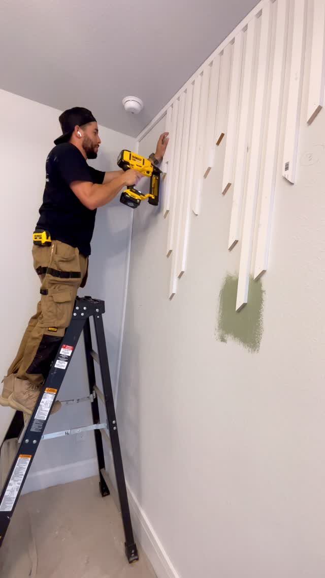 A man on a ladder uses a power drill to install white wooden slats on a wall.